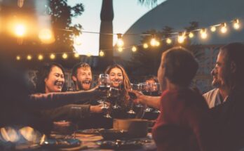 A group of young adults sits outside around a table with string lights above. They are clinking their wine glasses together.