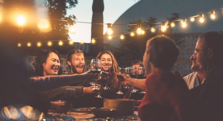 A group of young adults sits outside around a table with string lights above. They are clinking their wine glasses together.