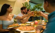 A group of friends gathering to share a large meal in a home. They are touching their beverage glasses together.