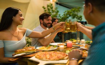A group of friends gathering to share a large meal in a home. They are touching their beverage glasses together.