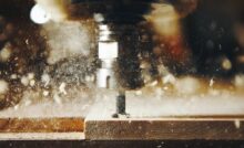 An active CNC machine bit tunneling a path through a flat piece of wood, with shavings flying around the surface.