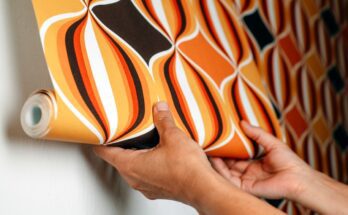 Close-up of a person's hands attaching wallpaper with a geometric designs and loud orange and brown colors to a wall.
