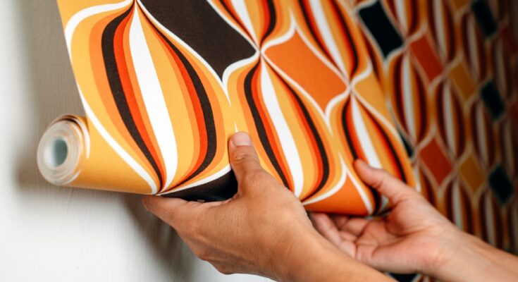 Close-up of a person's hands attaching wallpaper with a geometric designs and loud orange and brown colors to a wall.