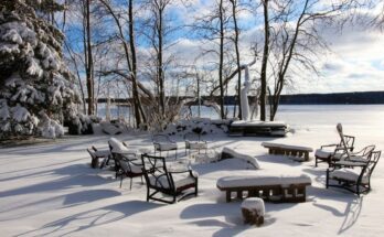 A backyard with patio furniture that is covered in a thick layer of snow. The yard overlooks a frozen pond.