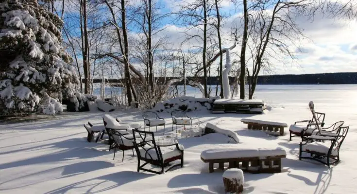 A backyard with patio furniture that is covered in a thick layer of snow. The yard overlooks a frozen pond.