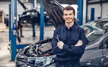 A smiling mechanic in a blue uniform holds a wrench while standing in front of a gray car in an auto shop.