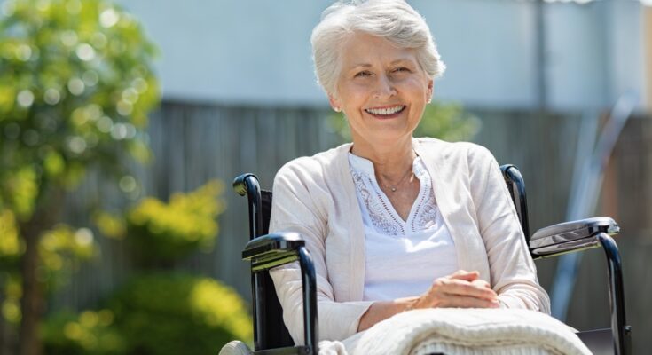 An older woman sitting outside in her wheelchair. She has a white blanket in her lap and is smiling.