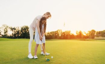 A woman shows her daughter how to hit a golf ball on a course by holding her hands on the club. Both are smiling.