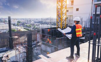 An engineer wearing a white hard hat holds a blueprint while standing on a ledge in a construction site.