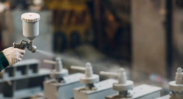A person in a protective glove uses a spray gun to coat metal components. The spray is a white powdery treatment.