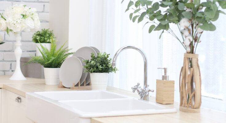 A cozy kitchen features a white sink with silver fixtures, a container of hand soap, and a vase with artificial flowers.