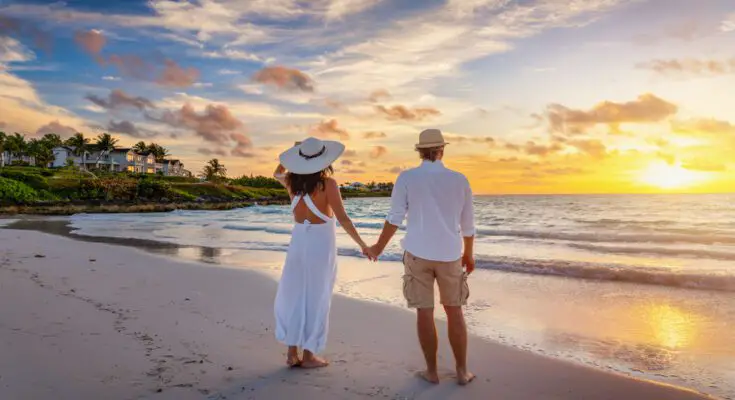 A man and a woman in summer clothes holds hands on a tropical beach while watching the sunset in the distance.