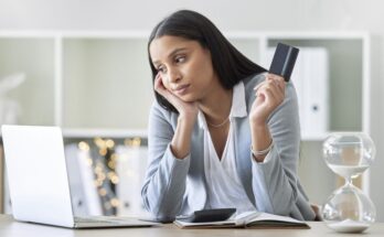 A woman in business casual attire holding a credit card while looking at a laptop on the table in front of her.