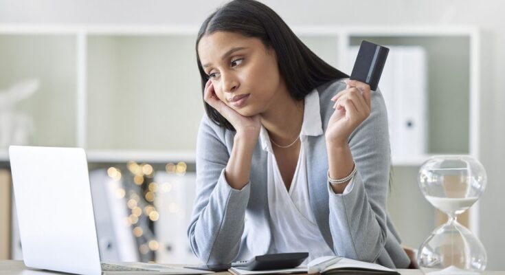 A woman in business casual attire holding a credit card while looking at a laptop on the table in front of her.