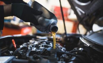 A person wearing black gloves pours oil from a black container into the oil fill opening of a vehicle engine.