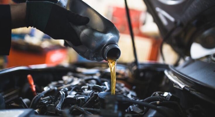 A person wearing black gloves pours oil from a black container into the oil fill opening of a vehicle engine.