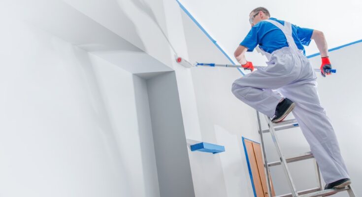 A man in a blue T-shirt and white overalls stands on a ladder while using a roller to paint the interior of a house.