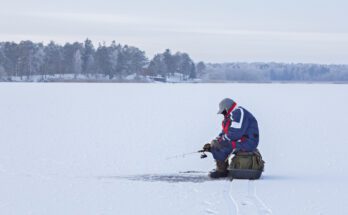 A man wearing heavy snow gear, sitting on a green bag placed on top of a frozen leg and holding a small fishing pole.