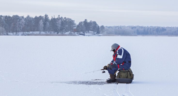 A man wearing heavy snow gear, sitting on a green bag placed on top of a frozen leg and holding a small fishing pole.