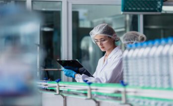A woman wearing protective eyewear and a hairnet holds a clipboard as she looks over water bottles in a factory.