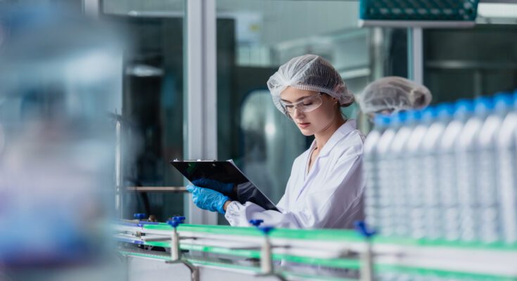A woman wearing protective eyewear and a hairnet holds a clipboard as she looks over water bottles in a factory.