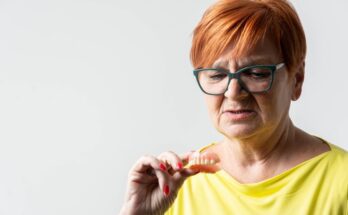 An older woman with red hair and glasses looks at the dentures she holds in her hand, examining them.