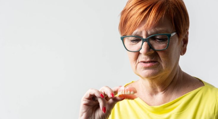 An older woman with red hair and glasses looks at the dentures she holds in her hand, examining them.