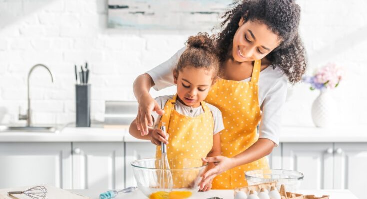 A mom and daughter wearing matching aprons stand in the kitchen as they use a whisk to beat eggs in a bowl.