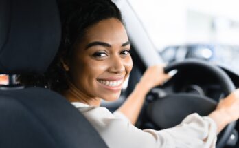 A woman looks over the shoulder of the driver's seat of a vehicle and smiles. She has her hands at 10 and 2 on the steering wheel.