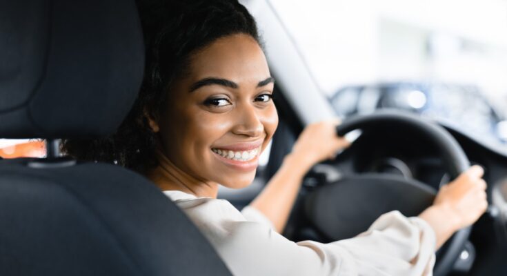 A woman looks over the shoulder of the driver's seat of a vehicle and smiles. She has her hands at 10 and 2 on the steering wheel.