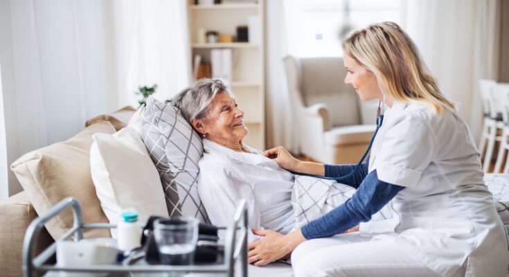 A senior woman lays in bed smiling at her provider as the provider uses a stethoscope. The walker is next to the bed.