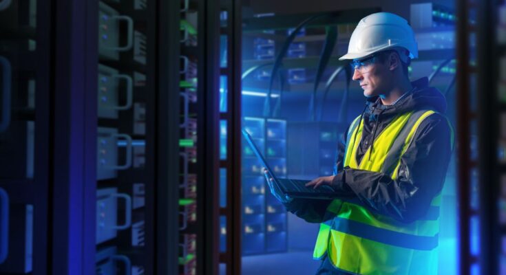 A man wearing a white hard hat and a yellow vest holds a laptop computer and stands in a data center aisle.