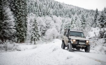 An off-road vehicle with a roof rack drives through a snowy forest trail surrounded by snow-covered pine trees in the winter.