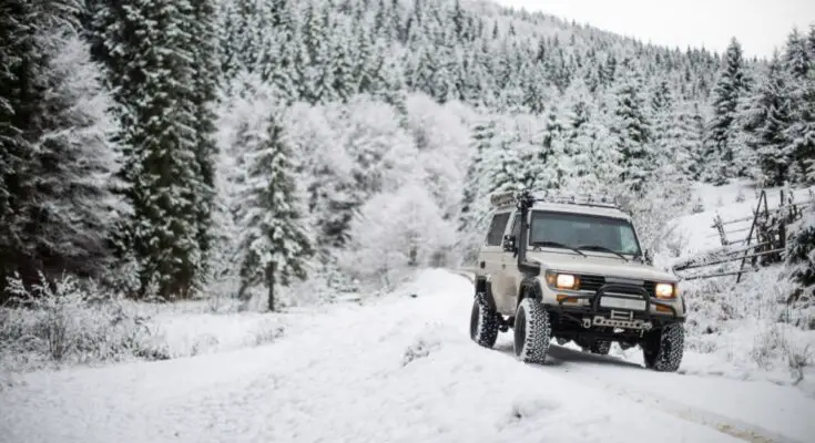 An off-road vehicle with a roof rack drives through a snowy forest trail surrounded by snow-covered pine trees in the winter.