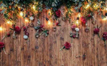 A dark wood backdrop for a photo booth is decorated with greenery, red and white flowers, and string lights.