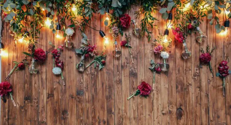 A dark wood backdrop for a photo booth is decorated with greenery, red and white flowers, and string lights.