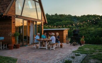 A group of people are sitting at a table outside. There is a house to the left of the frame and a dog by the table.