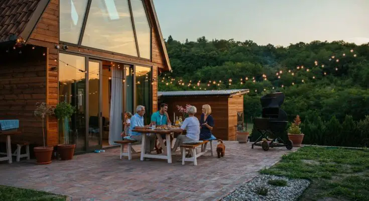 A group of people are sitting at a table outside. There is a house to the left of the frame and a dog by the table.