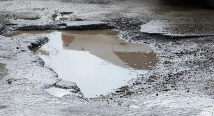 There is a giant pothole in asphalt. In the hole there is a puddle of murky standing water.