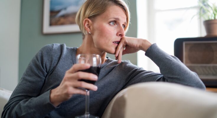 A blonde woman in a gray shirt sits on a couch, holding a glass of red wine with a worried expression.