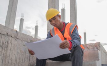 A male worker wearing a safety suit and helmet sits on a ladder and looks at the blueprint, and rests.