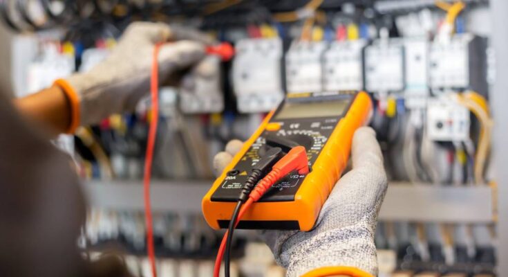 A person wearing gloves holds a multimeter. They're plugging in a wire into an electrical system control cabinet.