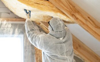 A person in a white jumpsuit is installing insulation into an attic. They are wearing protective glasses.