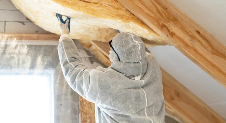 A person in a white jumpsuit is installing insulation into an attic. They are wearing protective glasses.