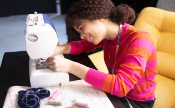 A teenager girl wearing a bright orange and pink sweater, sitting behind a desk, threading a white sewing machine.