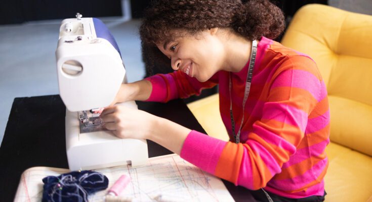 A teenager girl wearing a bright orange and pink sweater, sitting behind a desk, threading a white sewing machine.