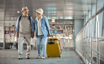 An elderly couple in matching outfits holds hands as they walk through an airport with their luggage behind them.
