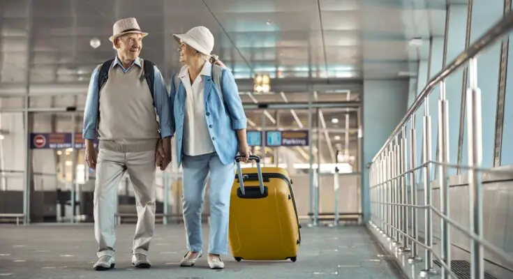 An elderly couple in matching outfits holds hands as they walk through an airport with their luggage behind them.