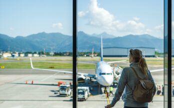 A woman wearing a backpack holds onto a piece of luggage while looking out the window of an airport.