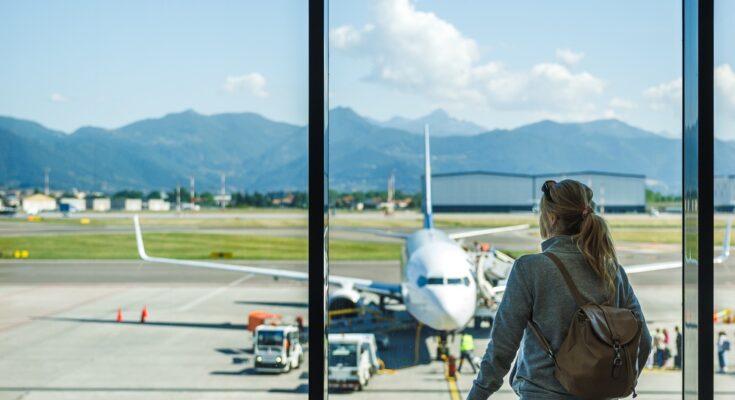 A woman wearing a backpack holds onto a piece of luggage while looking out the window of an airport.